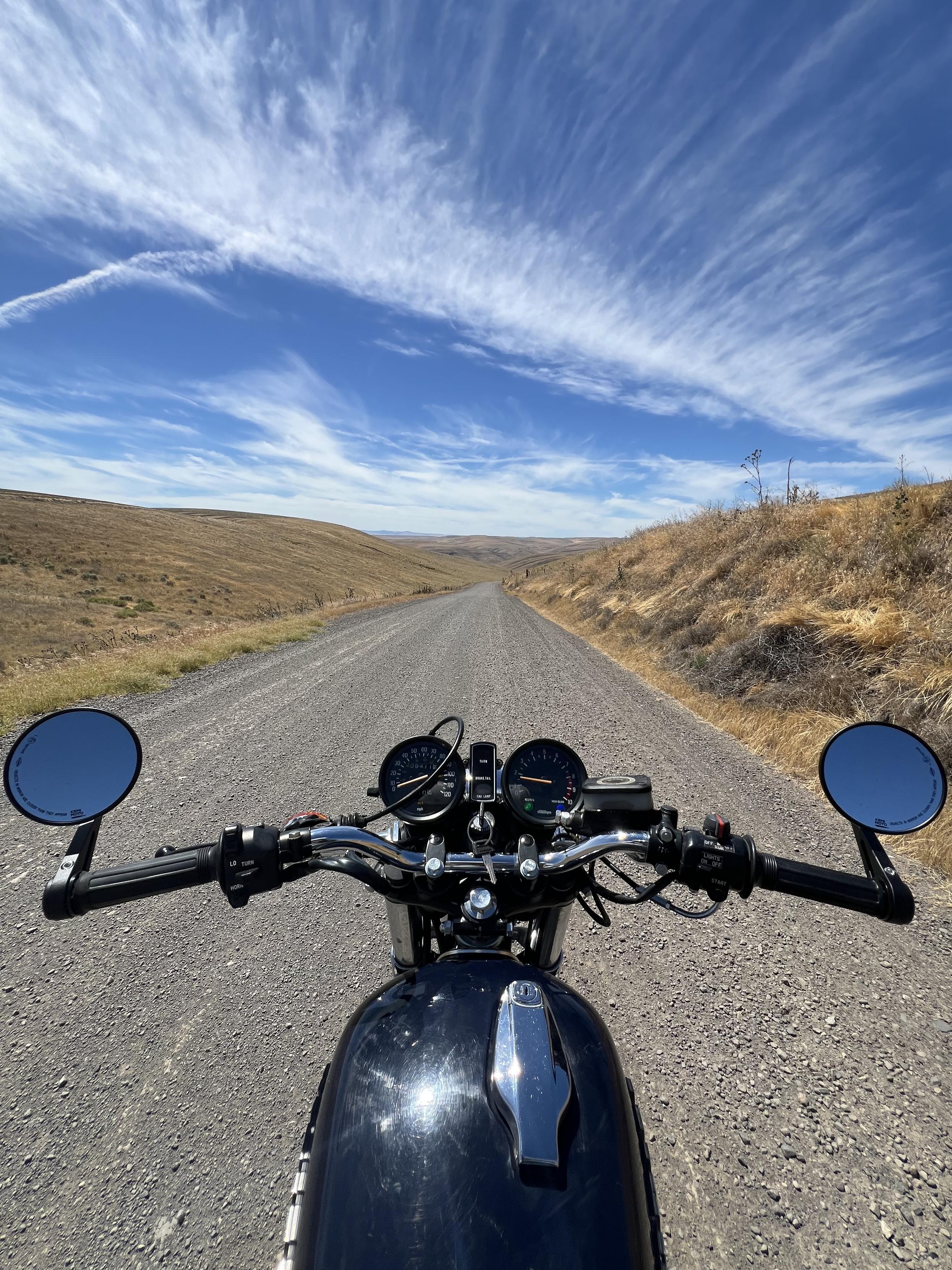 The view from the riding position on a motorcycle. The handlebars, tank and gauges are visible. Ahead a gravel road stretches out flanked by grassy brown verges. The sky has streaky white clouds.