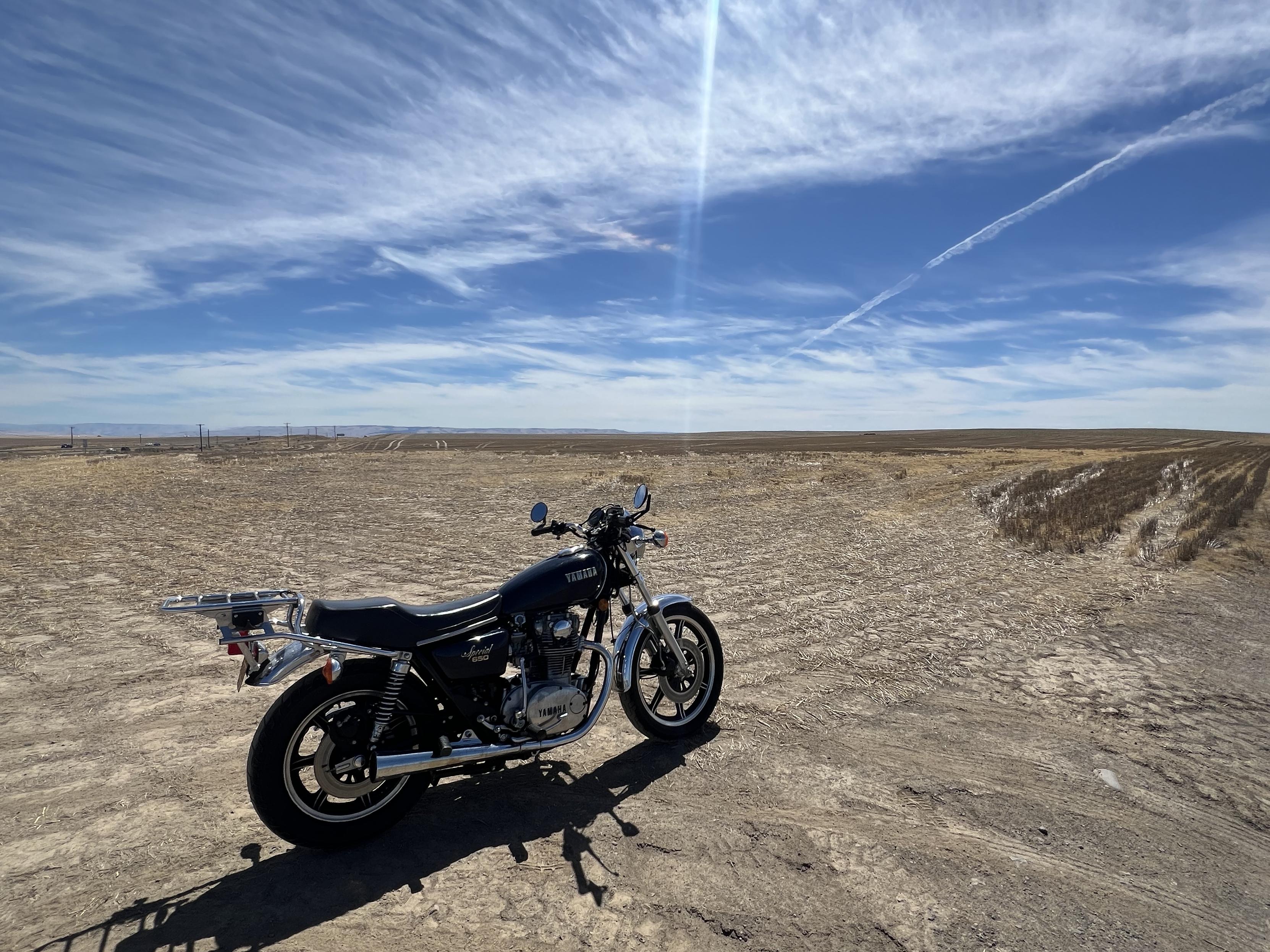 A blue motorcycle sits in an open brown field. The bike is facing to the right and the sun is shining off the chrome fender/mudguard.