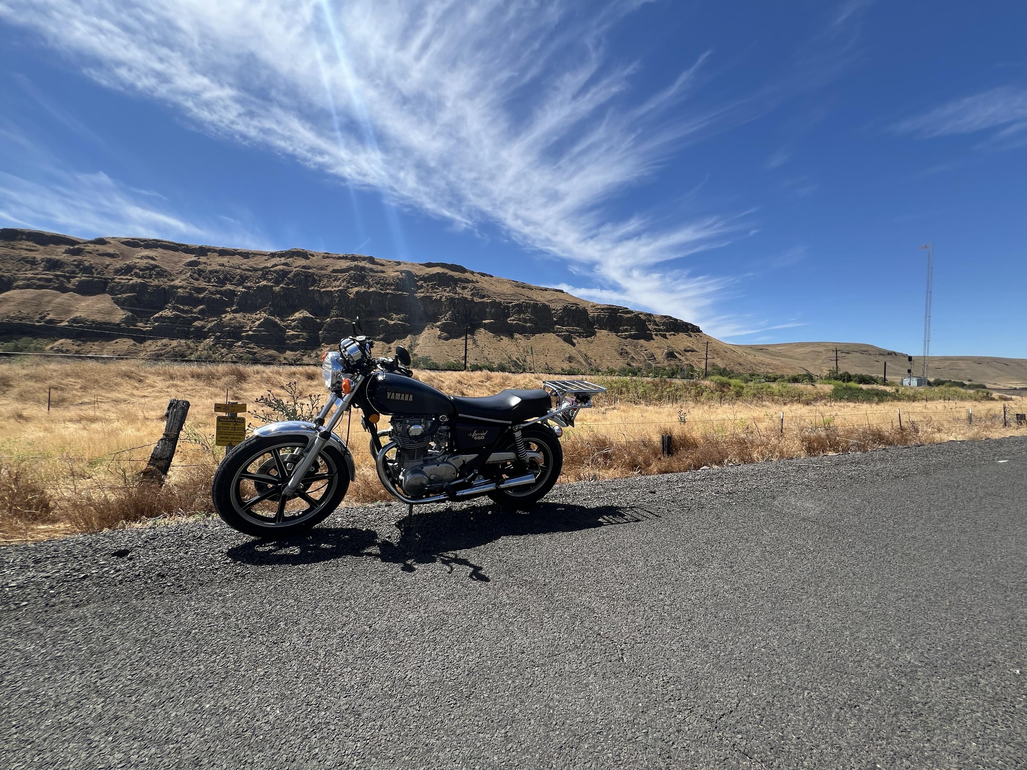 A blue motorcycle sitting at the side of the road. Behind the bike is a rocky outcrop. Visible to the right is an old radio antenna.