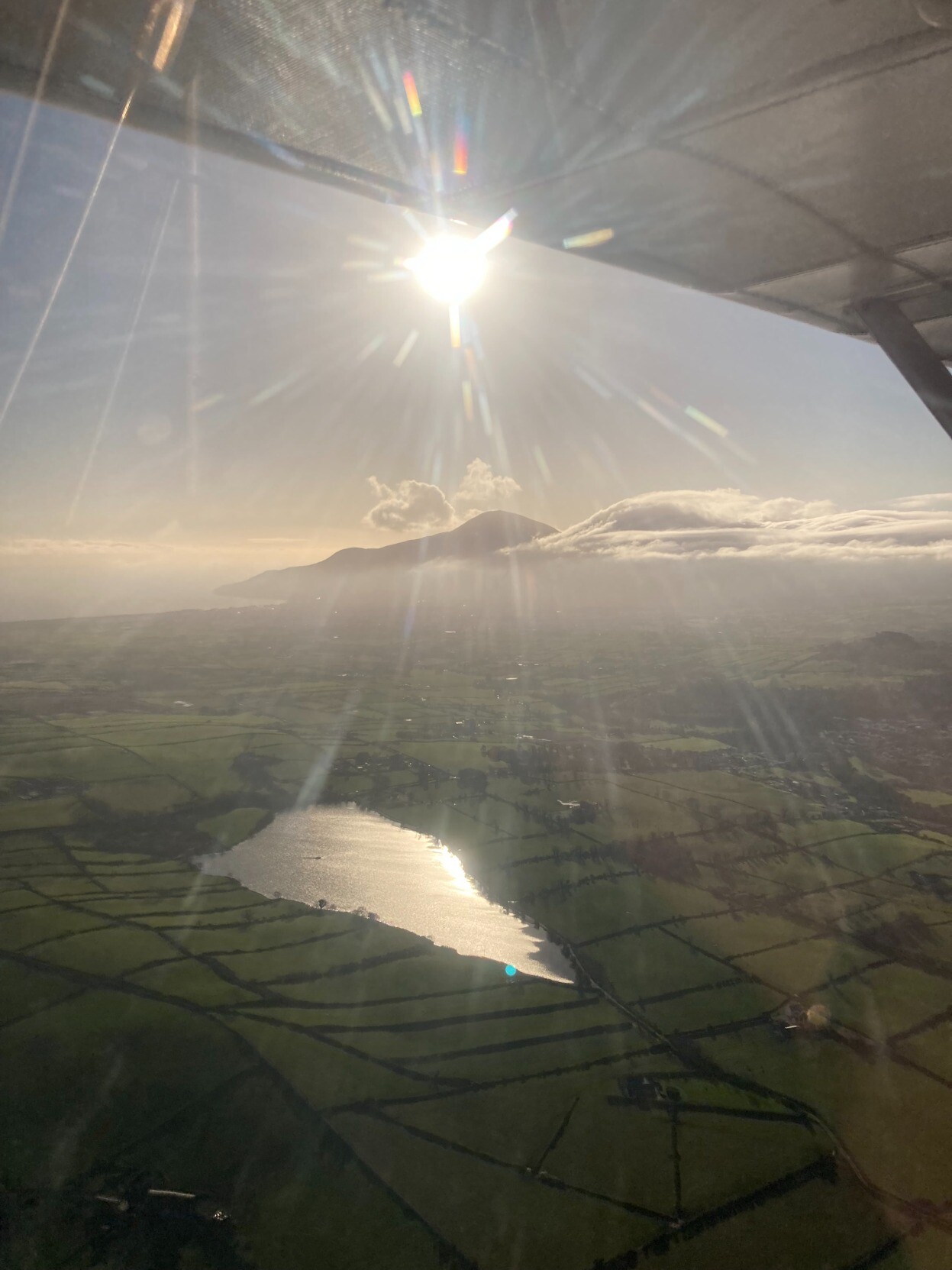 Slieve Donard in the distance with clouds rolling around it. I the foreground is a small lake surrounded by green fields. The wing of the plane the photo was taken from is visible in the top of the photo.