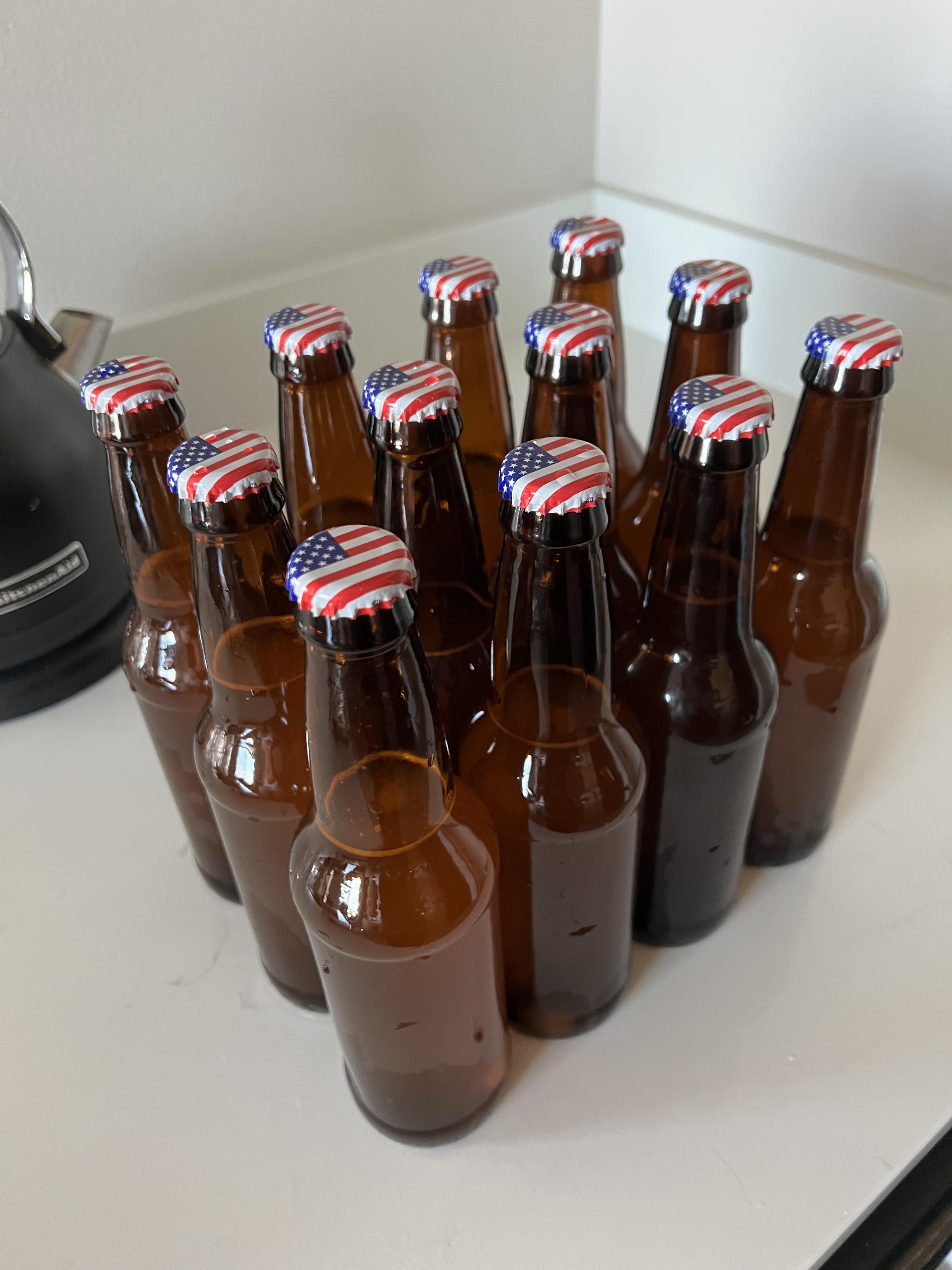 Sixteen brown glass bottles filled with beer. They are all aligned so that the American flag bottle caps are facing the same direction.